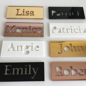 May include: Eight acrylic name plaques with different names cut out in the centre. The plaques are in various colours, including gold, black, white, and rose gold.