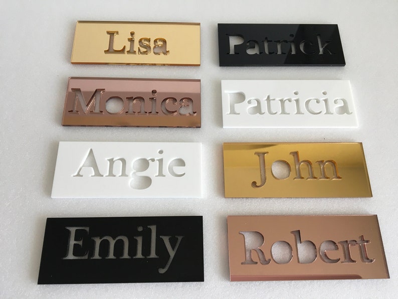 May include: Eight acrylic name plaques with different names cut out in the centre. The plaques are in various colours, including gold, black, white, and rose gold.