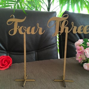 Puede incluir: Dos letreros de número de mesa de madera con las palabras "Cuatro" y "Tres" escritas en letra cursiva. Los letreros están sobre una mesa marrón con una rosa roja y flores rosas en el fondo.