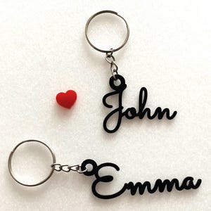 May include: Two black metal keychains with the names "John" and "Emma" written in cursive. The keychains are attached to silver key rings.