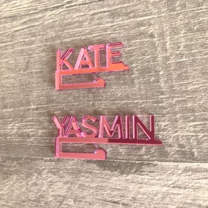 Two pink acrylic nameplates with a hook on the bottom. The top nameplate reads "KATE" and the bottom nameplate reads "YASMIN".