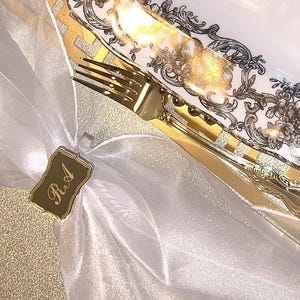 May include: A gold place setting with a white and gold patterned plate, a gold fork, and a gold place card with the name "Rita" written on it.