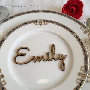 Peut inclure: Une d&eacute;coupe en bois en &eacute;criture cursive formant le pr&eacute;nom Emily, pos&eacute;e sur une assiette en c&eacute;ramique blanche avec un motif de bordure bleu, dor&eacute; et blanc. L'assiette est dispos&eacute;e sur une serviette beige &agrave; motifs avec une rose rouge.