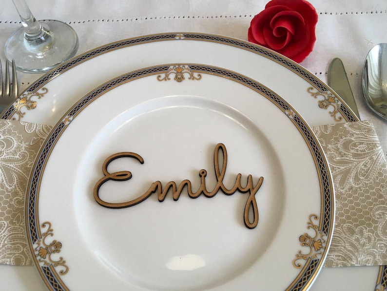 Peut inclure: Une d&eacute;coupe en bois en &eacute;criture cursive formant le pr&eacute;nom Emily, pos&eacute;e sur une assiette en c&eacute;ramique blanche avec un motif de bordure bleu, dor&eacute; et blanc. L'assiette est dispos&eacute;e sur une serviette beige &agrave; motifs avec une rose rouge.