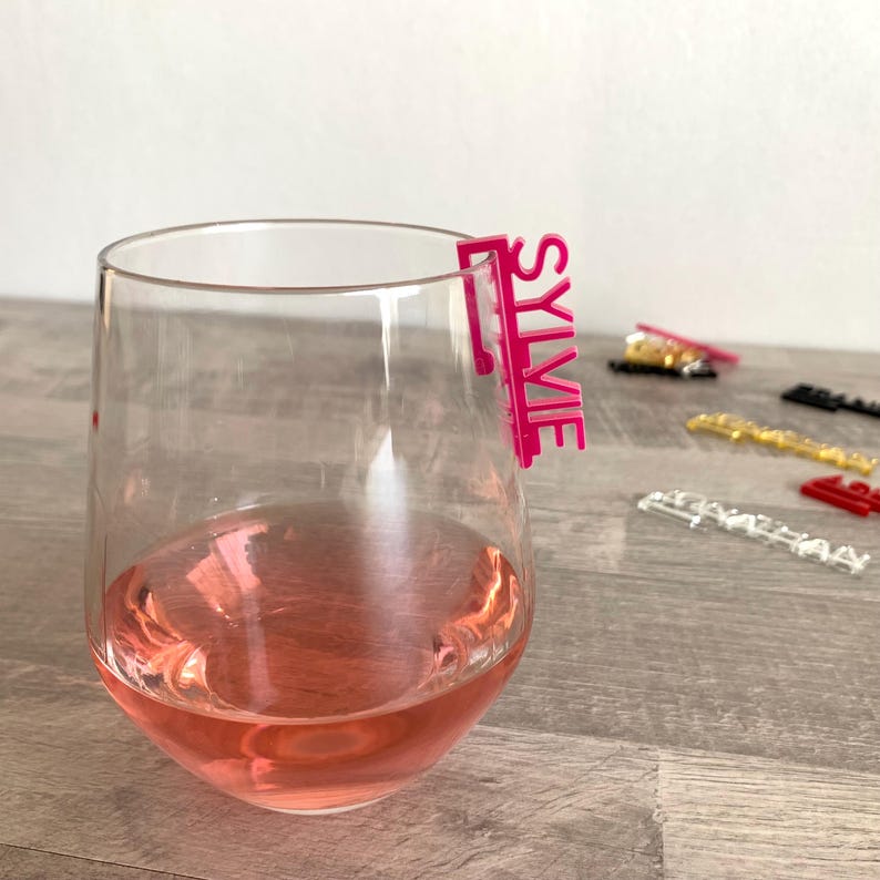 A pink acrylic glass marker with the name "Sylvie" written on it. The marker is attached to the rim of a glass of pink wine.