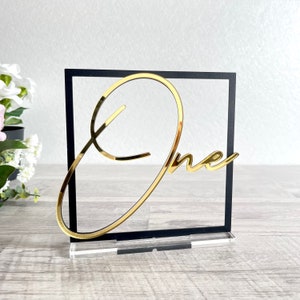 May include: Gold mirror acrylic 'one' table number with a black square frame and a clear acrylic base.
