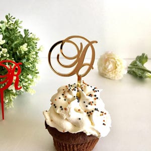 May include: A gold-coloured letter "D" cake topper on a cupcake with white frosting and sprinkles. The cupcake sits on a white surface, with a red letter "R" topper and greenery in the background.