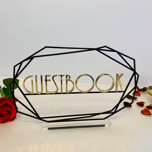 K&ouml;nnte beinhalten: Ein schwarzer geometrischer Rahmen mit einem goldenen Schild mit der Aufschrift "GUESTBOOK". Der Rahmen steht auf einem klaren Acrylsockel. Das Schild befindet sich auf einer wei&szlig;en Oberfl&auml;che mit einer roten Rose und Rosenbl&auml;ttern.