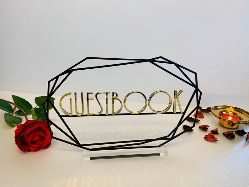 K&ouml;nnte beinhalten: Ein schwarzer geometrischer Rahmen mit einem goldenen Schild mit der Aufschrift "GUESTBOOK". Der Rahmen steht auf einem klaren Acrylsockel. Das Schild befindet sich auf einer wei&szlig;en Oberfl&auml;che mit einer roten Rose und Rosenbl&auml;ttern.