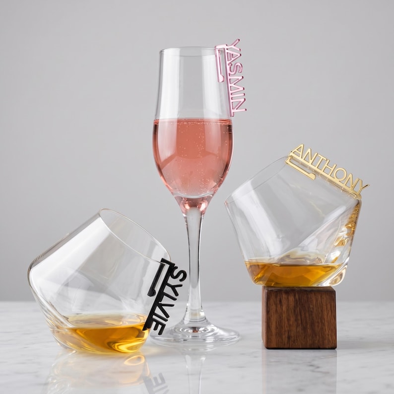 May include: Three glasses, including a champagne flute and two lowball glasses, are arranged on a white surface. The champagne flute holds a pink beverage and has a pink name tag that reads "YASMIN". The lowball glasses contain amber liquid and have name tags that read "SYLVIE" and "ANTHONY".