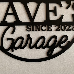 K&ouml;nnte beinhalten: Schwarzes Metallschild mit dem Text "Dave's Garage Since 2023" in einer abgerundeten, kursiv geschriebenen Schrift.