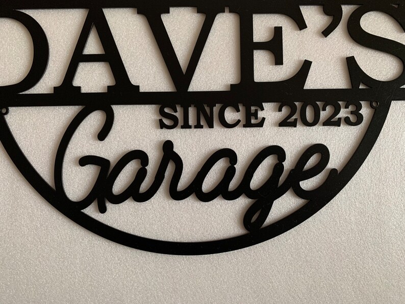 K&ouml;nnte beinhalten: Schwarzes Metallschild mit dem Text "Dave's Garage Since 2023" in einer abgerundeten, kursiv geschriebenen Schrift.