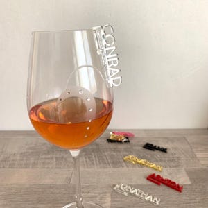 A clear glass wine glass with a light orange liquid inside. A clear acrylic wine glass charm with the name "CONRAD" is attached to the rim of the glass.