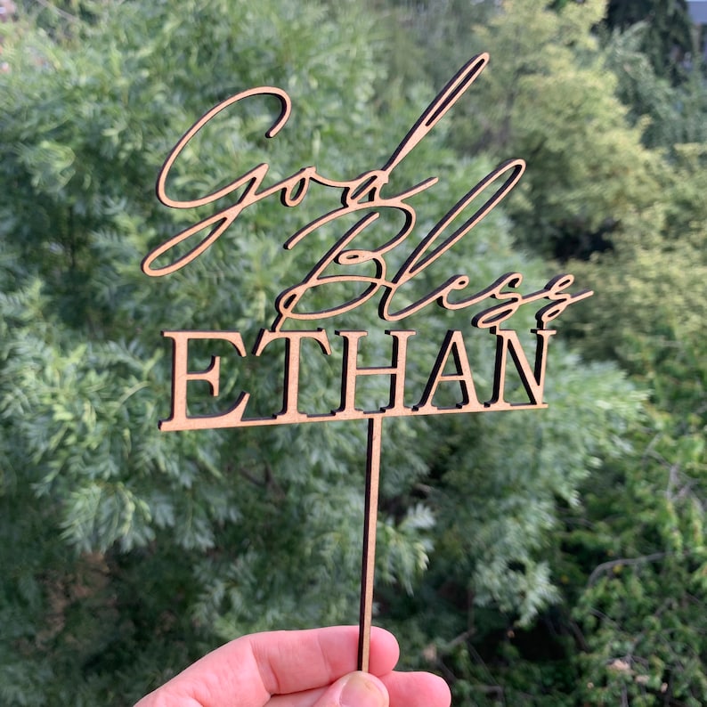 以下が含まれることがあります： 「God Bless Ethan」というテキストが書かれた木製のケーキトッパー。トッパーは茶色で、ケーキに差し込むための棒がついています。
