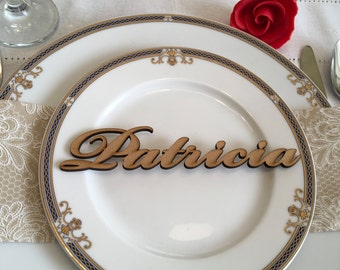 Puede incluir: Una tarjeta de lugar de madera con el nombre "Patricia" escrito en cursiva. La tarjeta de lugar está en un plato blanco con un borde dorado y azul.