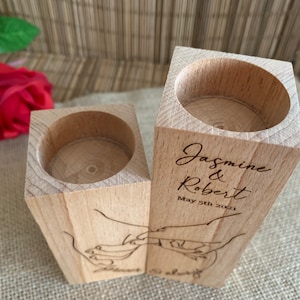 May include: Two wooden candle holders with a cutout design of hands holding each other. The holders are engraved with the names "Jasmine & Robert" and the date "May 5th 2021".