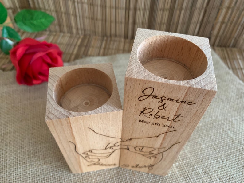 May include: Two wooden candle holders with a cutout design of hands holding each other. The holders are engraved with the names "Jasmine & Robert" and the date "May 5th 2021".