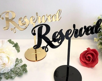 May include: Two acrylic table signs with the word "Reserved" in a cursive font. The sign on the left is gold and the sign on the right is black.