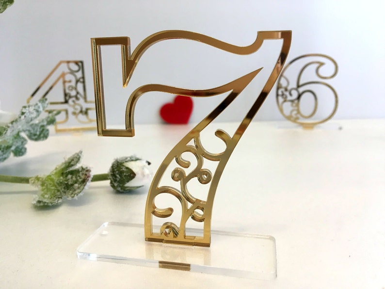 Pode incluir: N&uacute;mero de mesa sete em acr&iacute;lico dourado com um design floral decorativo. O n&uacute;mero est&aacute; em uma base de acr&iacute;lico transparente.