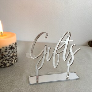 May include: A silver acrylic sign with the word "Gifts" written in a cursive font. The sign is on a mirrored base and is sitting on a white surface.