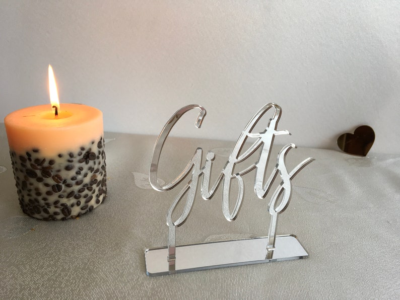 May include: A silver acrylic sign with the word "Gifts" written in a cursive font. The sign is on a mirrored base and is sitting on a white surface.
