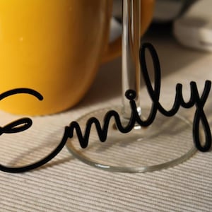 Peut inclure: Une &eacute;tiquette nominative en acrylique noir portant le pr&eacute;nom Emily &eacute;crit dans une police cursive. L'&eacute;tiquette est con&ccedil;ue pour &ecirc;tre fix&eacute;e &agrave; la base d'un verre &agrave; pied transparent, plac&eacute;e devant une tasse jaune vif.