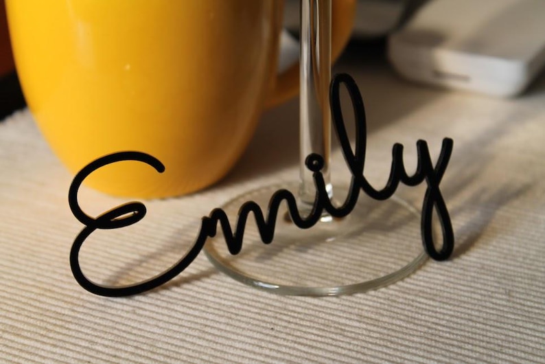 Peut inclure: Une &eacute;tiquette nominative en acrylique noir portant le pr&eacute;nom Emily &eacute;crit dans une police cursive. L'&eacute;tiquette est con&ccedil;ue pour &ecirc;tre fix&eacute;e &agrave; la base d'un verre &agrave; pied transparent, plac&eacute;e devant une tasse jaune vif.