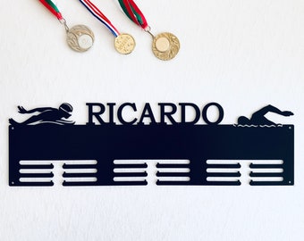 May include: Black metal medal display with the name "RICARDO" and swimmer silhouettes. The display has slots for medals and is hanging with three medals with red, white, and green ribbons.