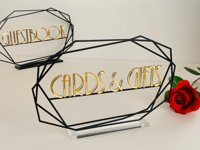 K&ouml;nnte beinhalten: Zwei schwarze geometrische Tischschilder mit goldenen Buchstaben. Das Schild links lautet "Guestbook" und das Schild rechts "Cards & Gifts".