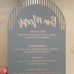 May include: A gray wooden sign with a gold arch design and the words "Bar Menu" written in gold script. The sign lists a variety of drinks, including Raspberry, Gin, and Cosmopolitans.
