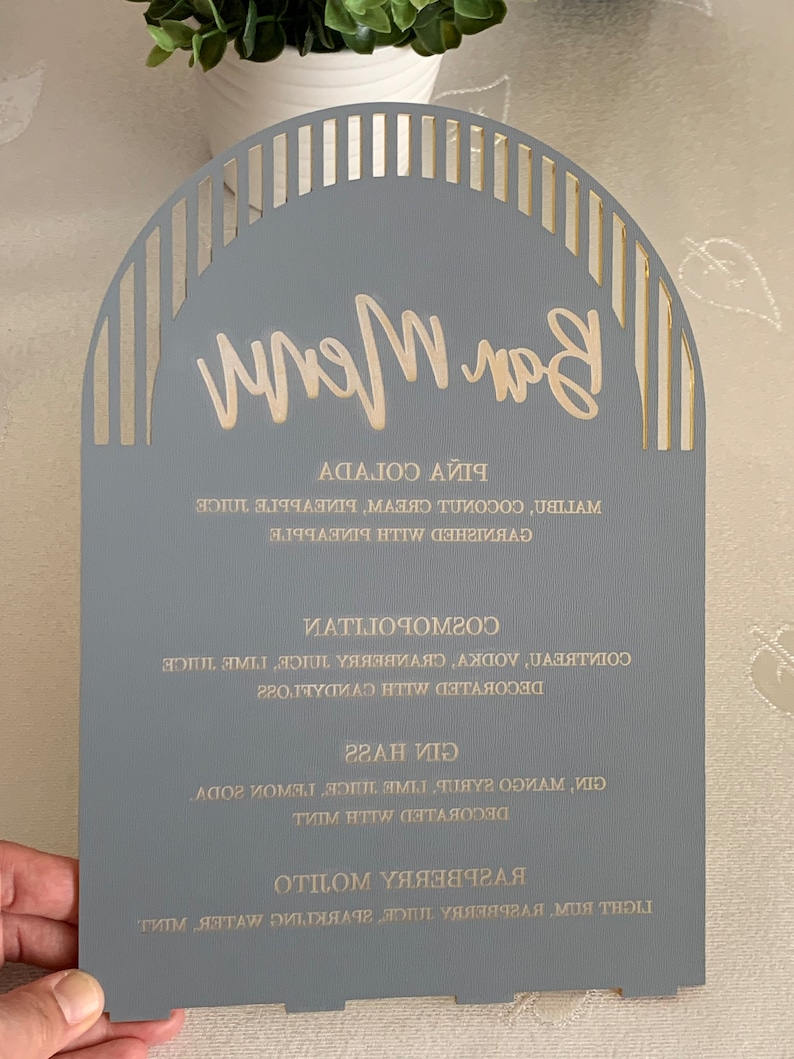 May include: A gray wooden sign with a gold arch design and the words "Bar Menu" written in gold script. The sign lists a variety of drinks, including Raspberry, Gin, and Cosmopolitans.
