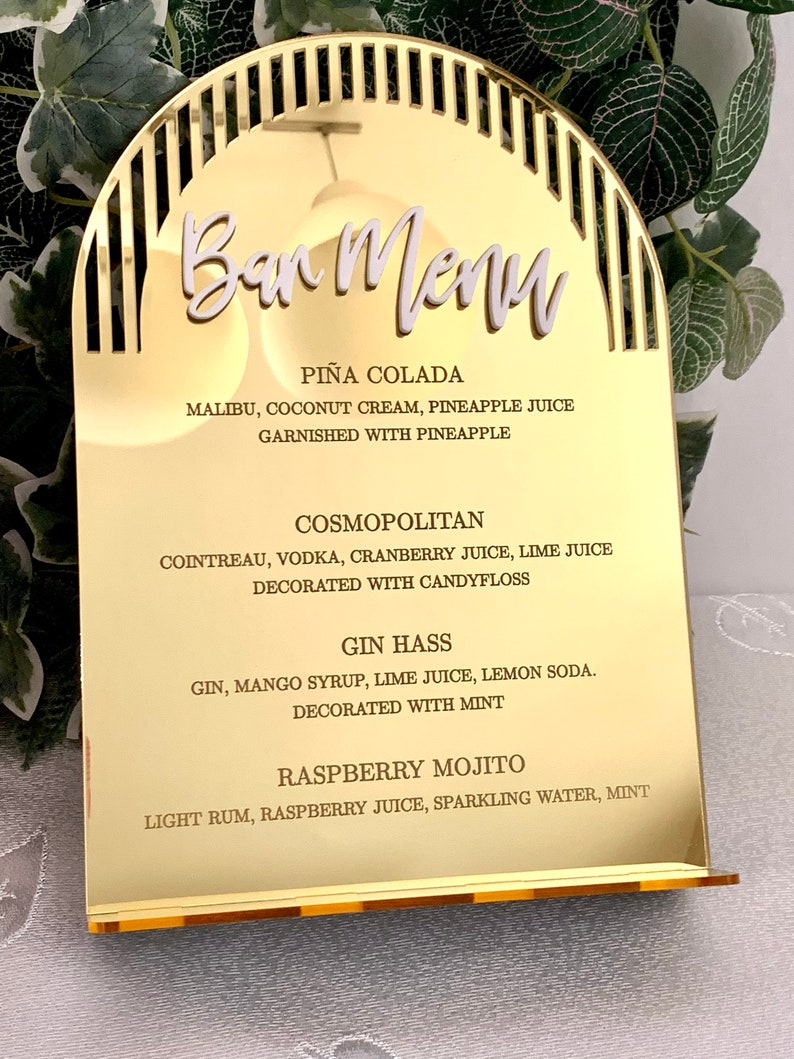 May include: Gold mirror bar menu with a decorative arch design. The menu lists four cocktails: Pi&ntilde;a Colada, Cosmopolitan, Gin Hass, and Raspberry Mojito. Each cocktail is described with its ingredients and garnishes.