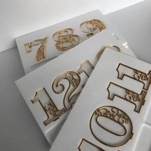 Pode incluir: Quatro n&uacute;meros de metal dourados, 7, 8, 9 e 10, com intrincados desenhos florais, s&atilde;o exibidos em placas de espuma branca.