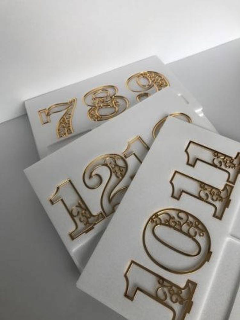 Pode incluir: Quatro n&uacute;meros de metal dourados, 7, 8, 9 e 10, com intrincados desenhos florais, s&atilde;o exibidos em placas de espuma branca.
