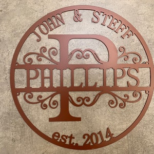 May include: A copper-colored metal family name sign. The sign is circular with the names "JOHN & STEFF" at the top and "PHILLIPS" in the center, with the year "est. 2014" at the bottom. The design includes decorative flourishes.