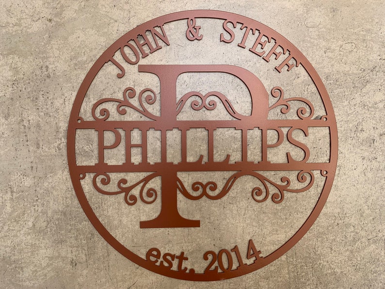 May include: A copper-colored metal family name sign. The sign is circular with the names "JOHN & STEFF" at the top and "PHILLIPS" in the center, with the year "est. 2014" at the bottom. The design includes decorative flourishes.