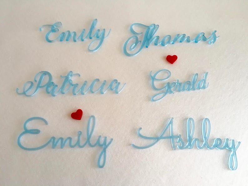 Peut inclure: Noms en acrylique bleu clair en &eacute;criture cursive lisibles comme Emily, Thomas, Patricia, Gerald, Emily et Ashley. Deux petits c&oelig;urs rouges sont plac&eacute;s entre les noms. Le lettrage pr&eacute;sente une finition brillante et lisse.