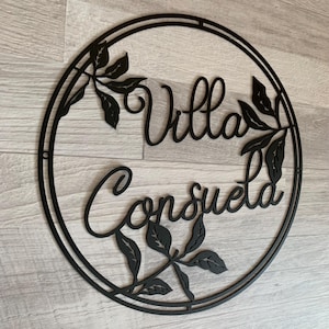 May include: Black metal sign with a circular frame and the words "Villa Consuelo" in a decorative script font. The sign is adorned with stylized leaf designs.