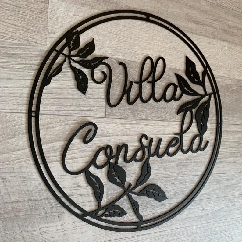May include: Black metal sign with a circular frame and the words "Villa Consuelo" in a decorative script font. The sign is adorned with stylized leaf designs.