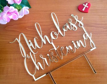 May include: Gold mirror acrylic cake topper with the text "Nicholas's Christening" in a cursive font. A small red heart with a silver cross is in the top right corner of the image.