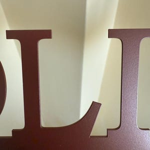May include: Close-up of a maroon metal sign with the letters "OL". The letters are cut out and have a slightly rounded font. The sign is against a light beige background.