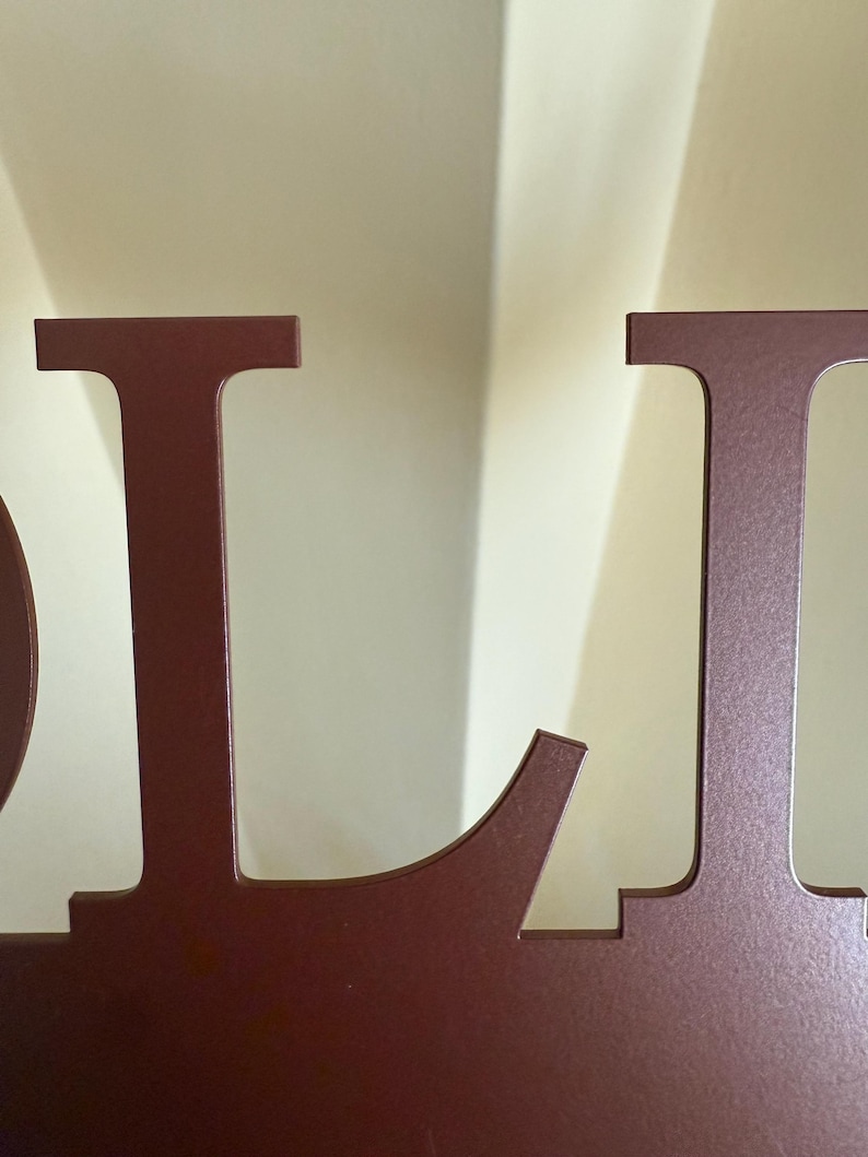 May include: Close-up of a maroon metal sign with the letters "OL". The letters are cut out and have a slightly rounded font. The sign is against a light beige background.