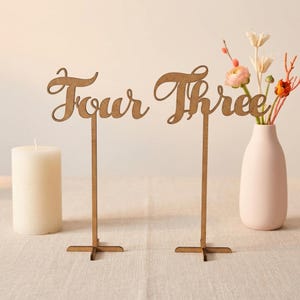May include: Two wooden table number signs, one reading "Four" and the other "Three", in a cursive font. A white candle and a pink vase with flowers are also present. The table numbers are made of light brown wood.