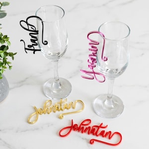 Puede incluir: Copas de champán con etiquetas de nombre personalizadas. Las etiquetas son en negro, rosa, dorado y rojo, con nombres como "Frank" e "Yarin" en fuente cursiva. Una pequeña planta verde en un jarrón gris está en el fondo.