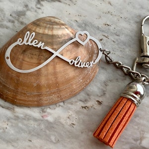May include: Silver infinity keychain with the names "ellen" and "oliver" engraved on it. The keychain has a heart charm and a tassel.