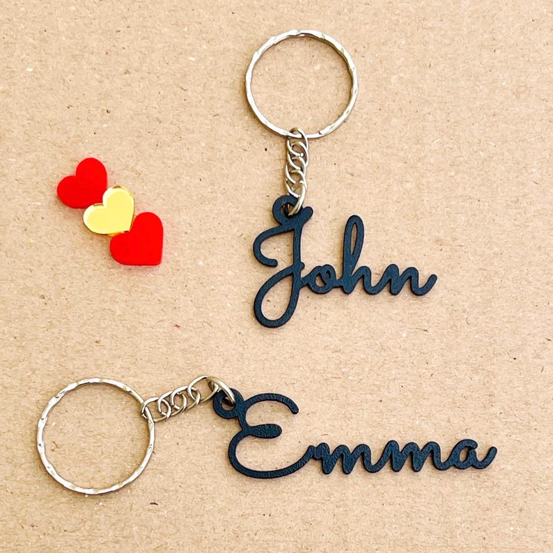 May include: Two black metal keychains with the names "John" and "Emma" written in a cursive font. The keychains are attached to silver key rings.