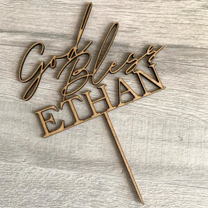 以下が含まれることがあります： 「God Bless Ethan」というテキストが書かれた木製のケーキトッパー。筆記体で書かれており、茶色で、ケーキに差し込むための棒がついています。