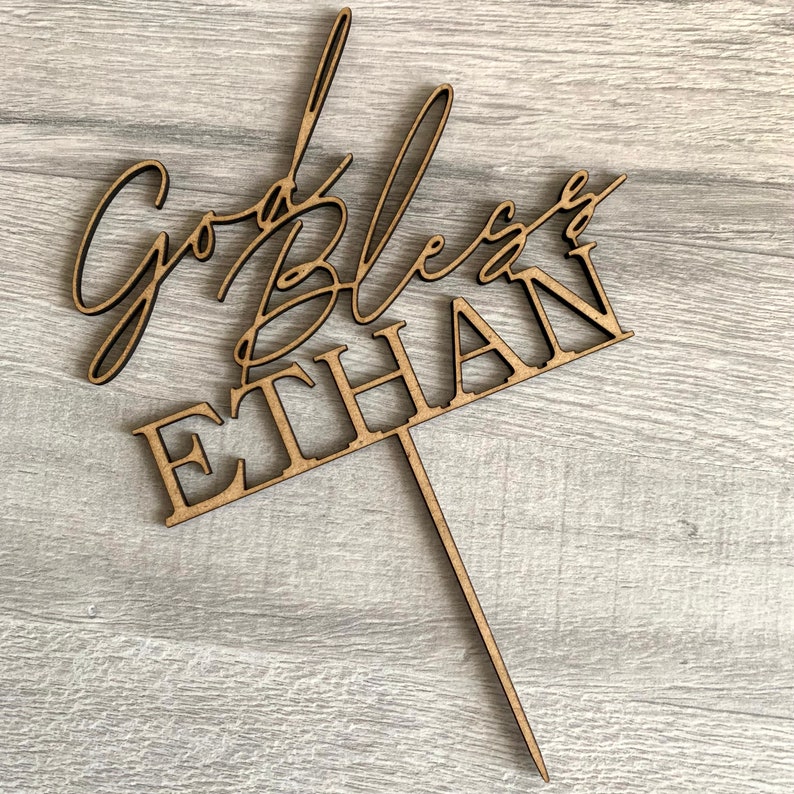 以下が含まれることがあります： 「God Bless Ethan」というテキストが書かれた木製のケーキトッパー。筆記体で書かれており、茶色で、ケーキに差し込むための棒がついています。