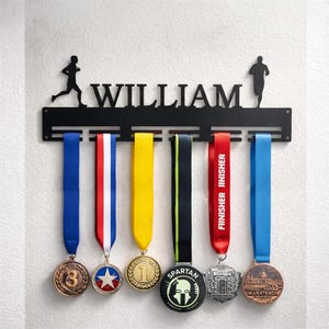 Könnte beinhalten: Ein schwarzer Medaillenhalter aus Metall zur Wandmontage mit dem Namen WILLIAM in Großbuchstaben, flankiert von zwei Läufer-Silhouetten. Die Halterung hat sechs horizontale Schlitze mit verschiedenen bunten Bändern und Medaillen.