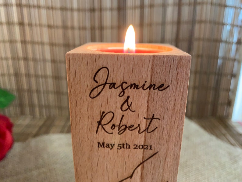 May include: A wooden candle holder with a lit candle inside. The holder is engraved with the names "Jasmine & Robert" and the date "May 5th 2021".